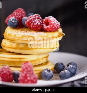 Pila di pancake con lamponi e mirtilli rivestite con zucchero a velo sulla piastra bianca e sfondo nero Foto Stock