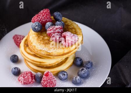 Pila di pancake con lamponi e mirtilli rivestite con zucchero a velo sulla piastra bianca e sfondo nero Foto Stock