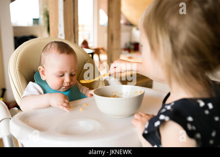 Irriconoscibile toddler girl a casa alimentando il suo bambino fratello. Foto Stock