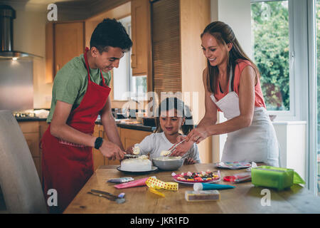 Madre decorare la torta con il figlio e la figlia nella cucina di casa propria. Essi coprono la torta con la glassa e avere caramelle per decorare. Foto Stock