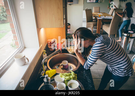 Ha sottolineato la mamma a casa. Ha la sua testa con le mani in un confuso lavello da cucina e i suoi figli sono rotondi in esecuzione in background. Foto Stock