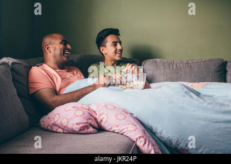 Padre e figlio di guardare un film insieme. Essi sono raggomitolati sul divano con popcorn e sono a ridere. Foto Stock