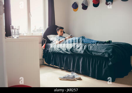 Ragazzo adolescente sdraiato sul suo letto di casa utilizzando un telefono intelligente. Foto Stock