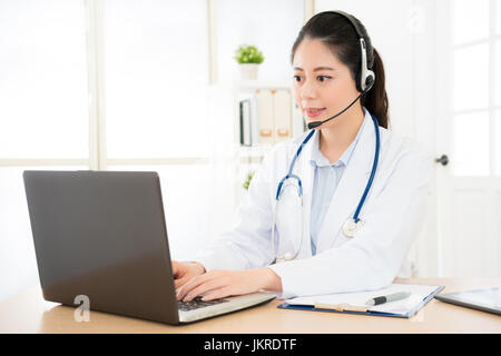 Medico donna attraverso internet conversazione mentre guardando il computer in ospedale con il paziente e la digitazione di tutti i record contenuti in un file dati di computer Foto Stock