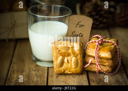 Pila di mandorla fatti in casa biscotti di Natale legato con rosso e nastro bianco, un bicchiere di latte, nota con parole scritte per babbo natale, in stile rustico, fest Foto Stock