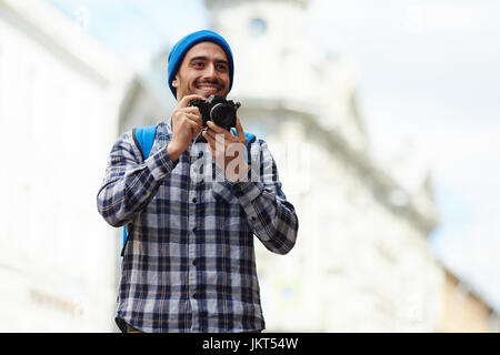 Ritratto del turista moderno sul viaggio Da solista in Europa, giovane uomo in piedi in strada di vecchia città di scattare foto con la fotocamera e sorridente Foto Stock