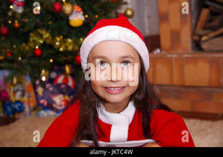 Bella poco sorridente ragazza indossando un abbigliamento di natale con un albero di Natale sfondo con alcuni regali Foto Stock