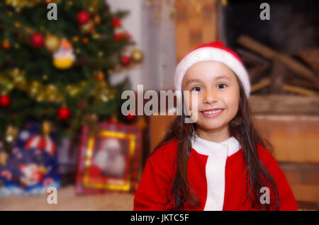 Bella poco sorridente ragazza indossando un abbigliamento di natale con un albero di Natale sfondo con alcuni regali Foto Stock