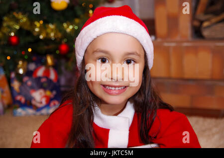 Bella poco sorridente ragazza indossando un abbigliamento di natale con un albero di Natale sfondo con alcuni regali Foto Stock