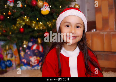 Bella poco sorridente ragazza indossando un abbigliamento di natale con un albero di Natale sfondo con alcuni regali Foto Stock