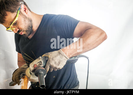 Un giovane uomo saldatore in una black T-shirt, occhiali di protezione e guanti di costruzione metallica elabora una smerigliatrice angolare su bianco backgroun isolato Foto Stock