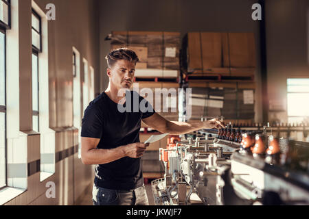 Il birraio esaminando la produzione di birra birreria in pianta. Giovane uomo in piedi alla birra macchina imbottigliatrice in fabbrica. Foto Stock