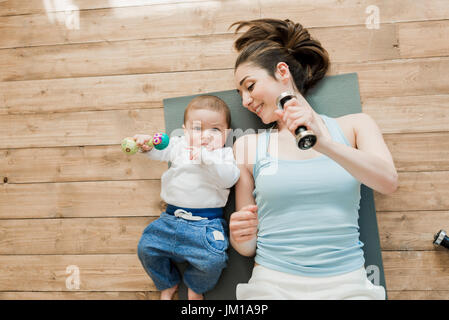 Vista superiore della madre con bambino sdraiato sul pavimento e giocando con manubri Foto Stock