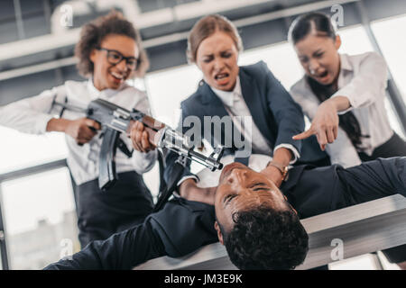 Tre giovani imprenditrici arrabbiato con un fucile punire l uomo d affari sdraiato sul tavolo, riunioni di team aziendali concetto Foto Stock