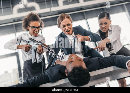 Tre giovani imprenditrici arrabbiato con un fucile punire l uomo d affari sdraiato sul tavolo, riunioni di team aziendali concetto Foto Stock