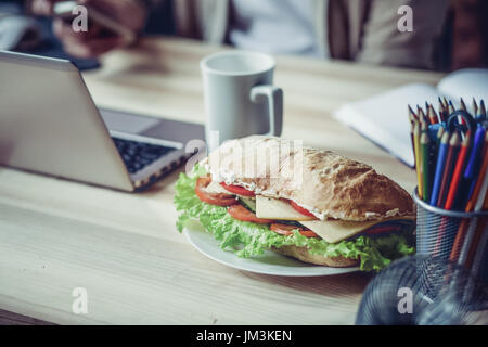 Vista ravvicinata di lavorare palce di freelancer durante il pranzo. Scrivania in legno con il computer portatile, una tazza di caffè e sandwich su di esso. Foto Stock