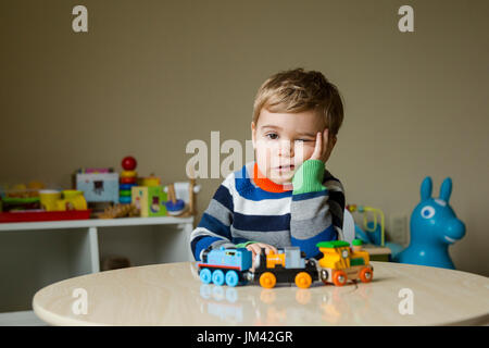 Un ragazzo bambino seduto nella sua sala giochi si appoggia sul suo i gomiti su un tavolo e squishes le sue guance con uno sguardo di tristezza sul suo volto. Foto Stock