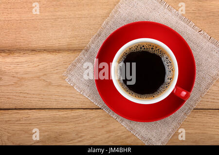 Piena grande tazza di nero caffè istantaneo con schiuma sul rosso piattino porcellana su tavola di legno con tovaglia tessile igienico, close up, elevati vista dall'alto, Foto Stock
