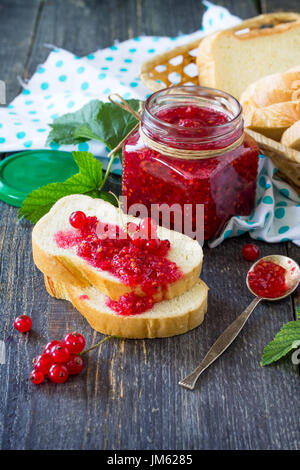 Marmellate fatte in casa. Un vaso di vetro con red confettura di ribes e pane bianco su una cucina un tavolo di legno. Conserve di Berry. Foto Stock