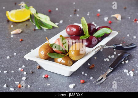 Olive nere e verdi in una piastra bianca su un grigio Sfondo astratto con limone, aglio e spezie. Cucina mediterranea. Mangiare sano concetto. Foto Stock