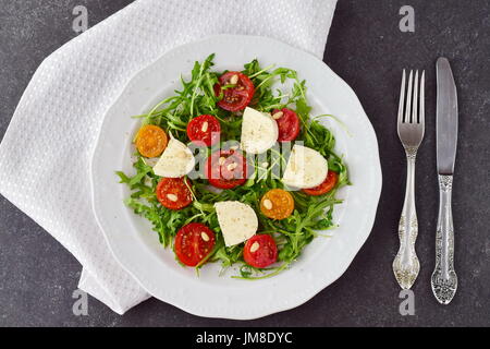 Tradizionale Italiano con insalata di pomodoro ciliegino, rucola, mozzarella, olio di oliva aceto di vino su una piastra bianca su un grigio Sfondo astratto Foto Stock