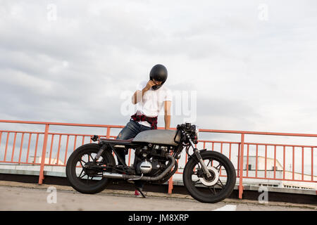 Abbastanza felice cavaliere uomo con barba e baffi decollare black moto casco e sedersi sul suo stile classico biker cafe racer moto. Moto custom made ho Foto Stock
