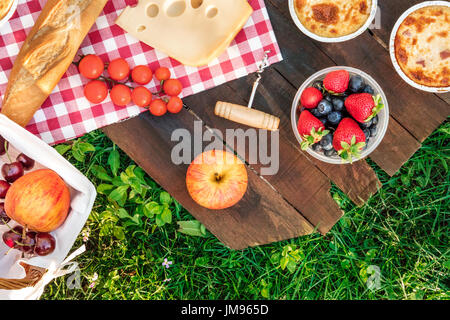 Foto aerea del picnic con mele, frutta fresca in contenitore di plastica e un pezzo di formaggio Olandese, quiche, baguette e un cavatappi, su di un legno rustico Foto Stock