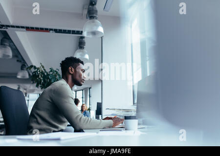 Vista laterale del giovane africano che lavorano in ufficio. Executive maschile seduto alla sua scrivania e di lavoro sul computer desktop. Foto Stock