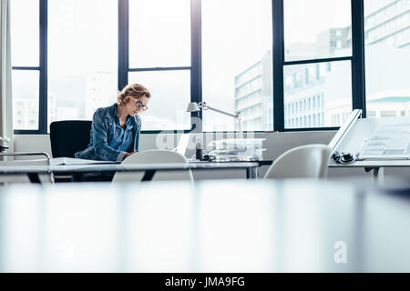 Donna manager seduto alla sua scrivania e di lavoro sul computer portatile. Bella giovane donna in cerca di lavoro occupato sulla scatola di piani di progetto. Foto Stock