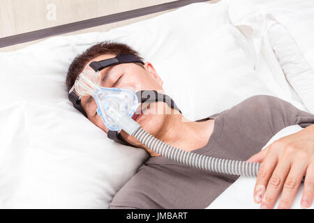 La mezza età uomo asiatico dormire nel suo letto INDOSSARE COPRICAPO CPAP maschera collegata al tubo flessibile dell'aria, dispositivo per persone con apnea nel sonno Foto Stock
