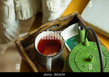 Off-bianco felpa lavorata a maglia che pende sulla sedia in legno, la tazza con il frutto rosso tè, pot nel vassoio da finestra, vecchio libro autunno autunno umore, accogliente Foto Stock