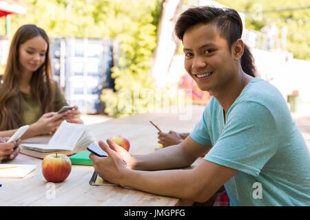 Foto di sorridente multietnica giovani amici studenti esterni utilizzando i telefoni cellulari. Uomo asiatico guardando la fotocamera. Foto Stock