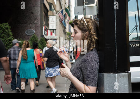 Giovane donna con capelli ricci, parlando al telefono utilizzando le cuffie / auricolari, tenendo il pezzo del microfono in mano mentre in piedi sulla strada Foto Stock