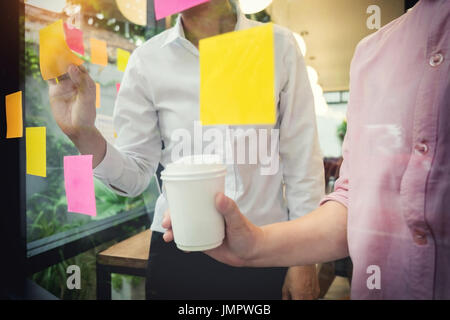 Uomo d'affari di incollaggio note adesive sulla parete di vetro in ufficio e discutere con il team. Foto Stock