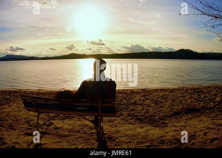 Da solo giovane in Silhouette seduti al sole Tourist prendere un periodo di riposo sul banco di legno al lago d'autunno. Foto Stock