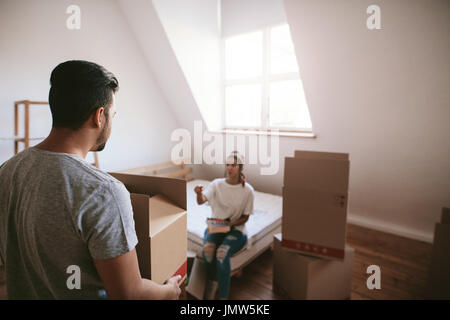 Vista posteriore del tiro del giovane uomo che porta cassetta con donna seduta sul letto in background. Coppia giovane in movimento nella nuova casa. Foto Stock