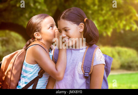 Studente con zaino whispering in orecchio amico contro alberi e prato nel parco Foto Stock