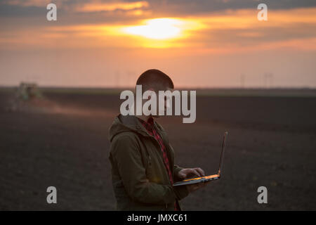 Giovane agricoltore con il computer portatile in piedi in campo con il trattore erpicatura in background Foto Stock