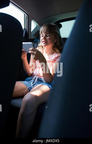 Felice ragazza adolescente in cuffie utilizzando il telefono cellulare nel sedile posteriore della macchina Foto Stock