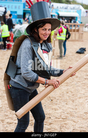 Cardboardia evento su Ramsgate Beach. Donna, 30s, nella stufa black hat, tenendo lungo palo di cartone, la testa girata a guardare al visualizzatore, sorridente, contatto visivo. Foto Stock