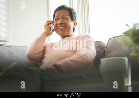 Donna sorridente tenendo il telefono mentre è seduto sul divano di casa Foto Stock