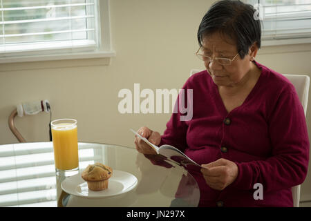 Senior donna libro lettura mentre seduti a tavola a casa Foto Stock