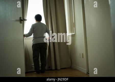 Vista posteriore del senior donna in piedi dalla finestra a casa Foto Stock