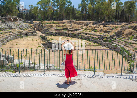 Concetto di viaggio turistico donna, vista di una giovane donna in un vestito rosso, scattando una foto delle rovine romane nel Parco Archeologico di Siracusa, Sicilia. Foto Stock