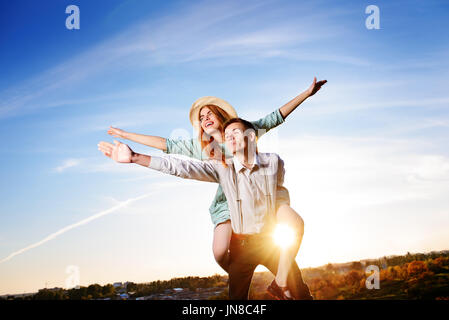 Giovane ragazzo piggybacking allegro ragazza come aereo sullo sfondo del cielo. Foto Stock