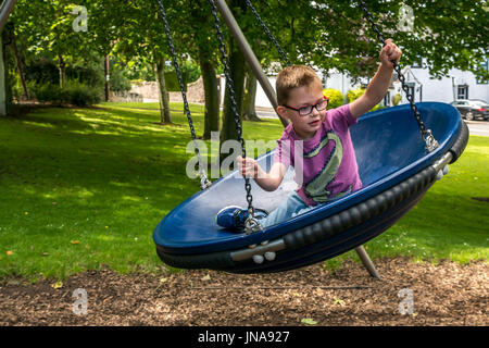 Chiusura del giovane ragazzo divertirsi seduti in oscillazione circolare nel parco giochi sul villaggio verde, Dirleton, East Lothian, Scozia, Regno Unito Foto Stock