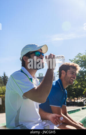 Due giocatori professionisti idratare dopo un duro gioco di tennis. Essi sono gli amici e sono seduti in appoggio per il gioco. Sono felici e ridere. Foto Stock