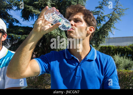 Giocatore di tennis professionista indossare una polo blu è in appoggio match di tennis di bere acqua da una bottiglia. Lui è seduto di fronte al campo da tennis. Foto Stock