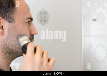 Uomo barba di trimming in bagno con rasoio elettrico sullo specchio Foto Stock