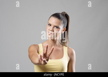 Stupendo giovane sportivo ritratto femminile che mostra due dita di pace segno a mano su grigio di sfondo per studio. Foto Stock